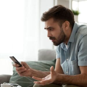 Photo of a confused looking man sitting in a living room, looking at his mobile phone.
