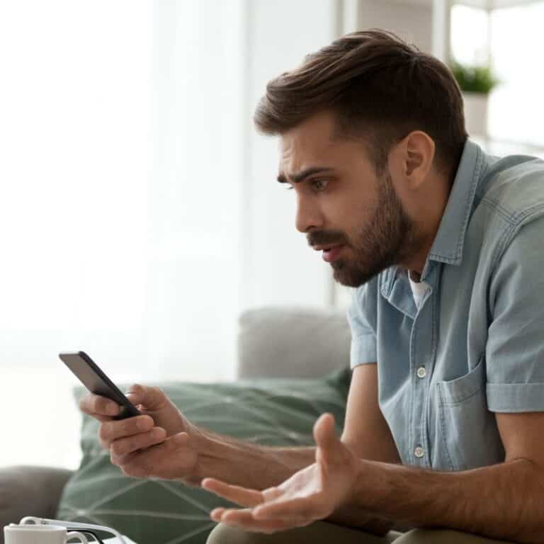 Photo of a confused looking man sitting in a living room, looking at his mobile phone.