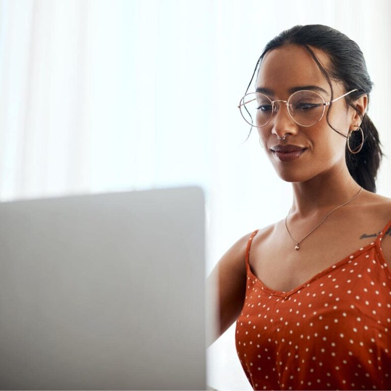 Photo of black woman with an orange shirt and glasses sitting at their computer. updating their wordpress plugins