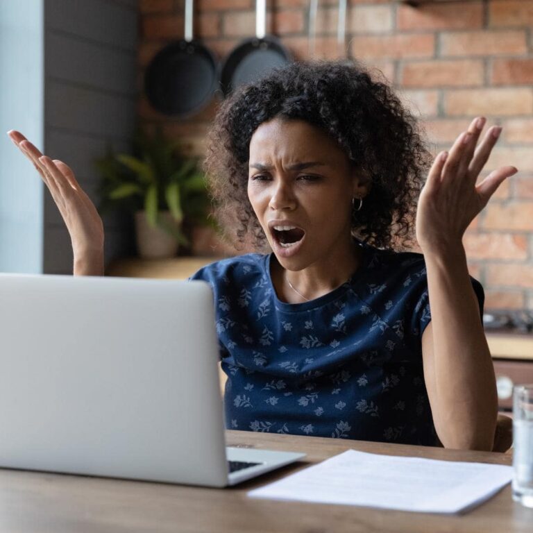 Photo of black woman sitting at her laptop, She looks frustrated with her computer because of slow load times.